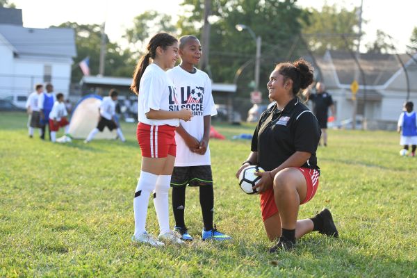 A female coach kneels on one knee while holding a soccer ball, smiling and talking with two young players—a girl and a boy—in white U.S. Soccer Foundation shirts. They stand on a sunny field with other players and equipment visible in the distance.